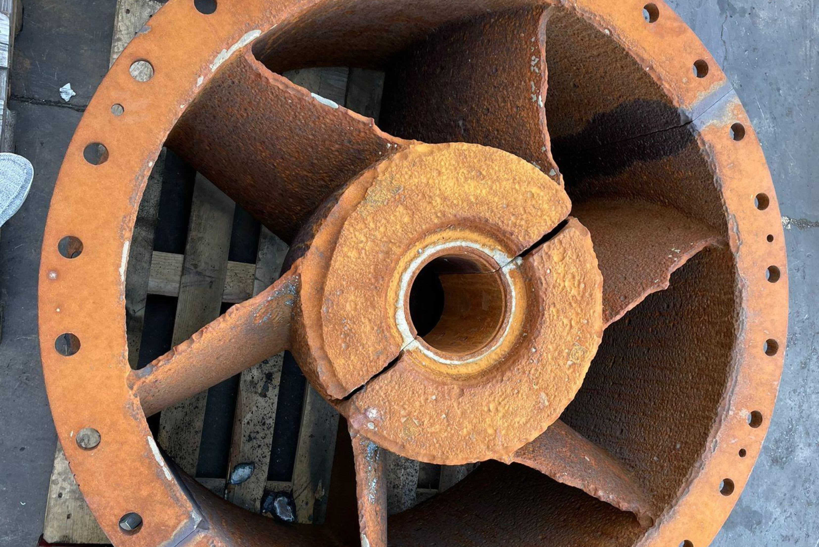 Un'unità di girante industriale o un rotore fortemente arrugginito, dotato di una piastra flangiata anulare e pale disposte radialmente, posizionato su un pallet di legno.