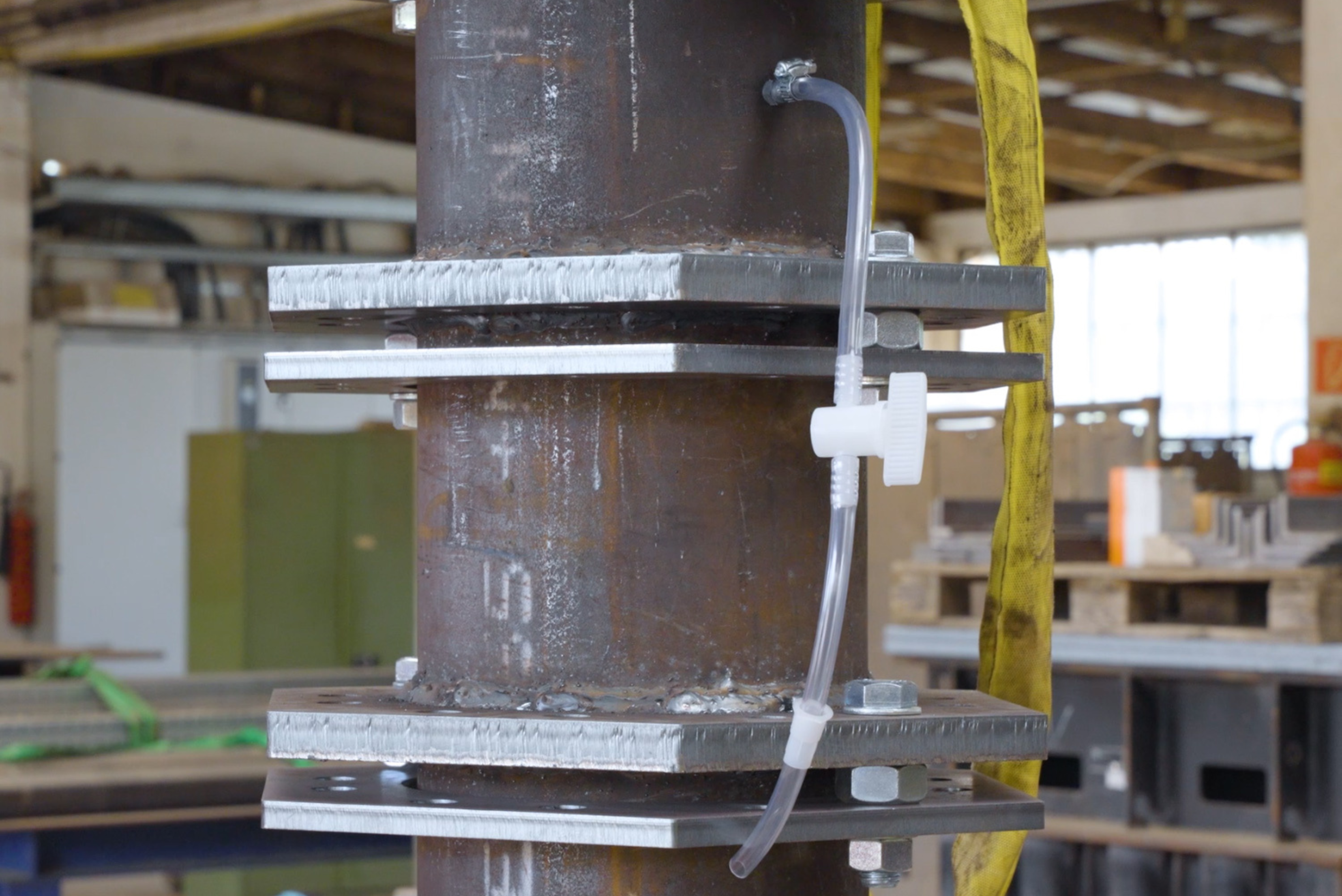 Un tubo metallico verticale e robusto, dotato di flange saldate e imbullonate, si trova all'interno di un'officina, con un tubicino trasparente e una valvola bianca collegati a un'apertura laterale.