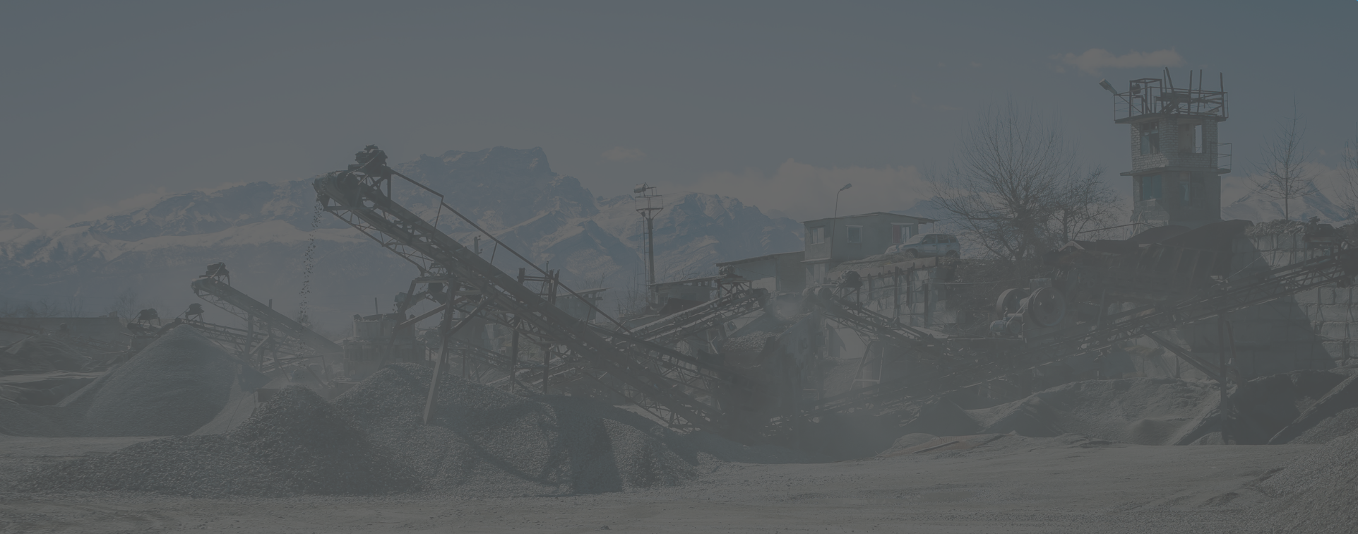 Panoramica di un impianto industriale con nastri trasportatori per inerti e cumuli di ghiaia, con montagne sullo sfondo sotto una foschia polverosa.