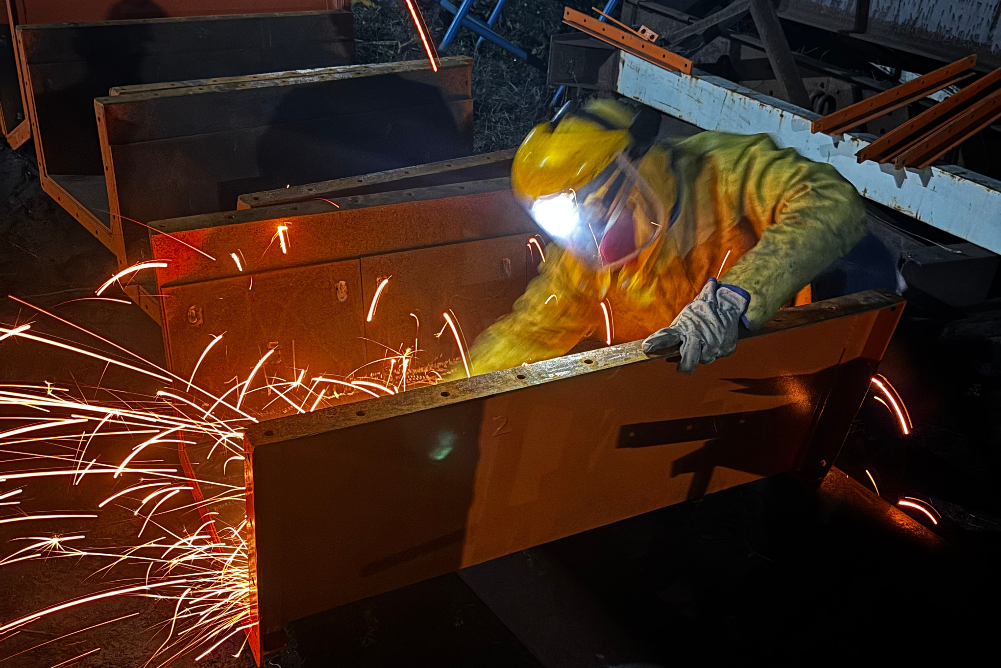 Un lavoratore indossa una tuta protettiva, guanti spessi e una visiera paraschizzi mentre lavora su componenti metallici, generando una pioggia di scintille arancioni brillanti.