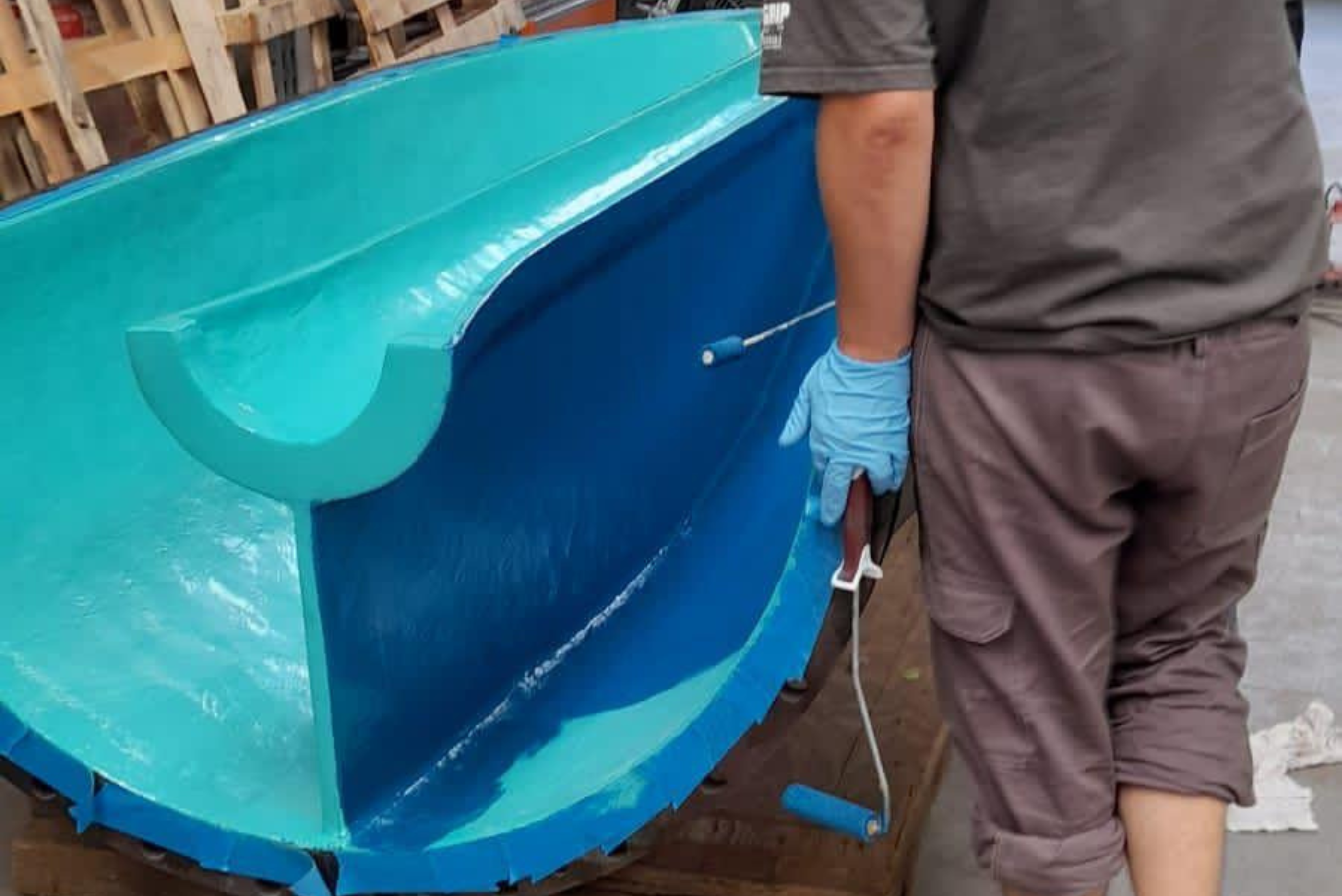 Un tecnico in abiti da lavoro e guanti protettivi azzurri applica un rivestimento protettivo blu all'interno di un grande alloggiamento industriale utilizzando un rullo per verniciatura.