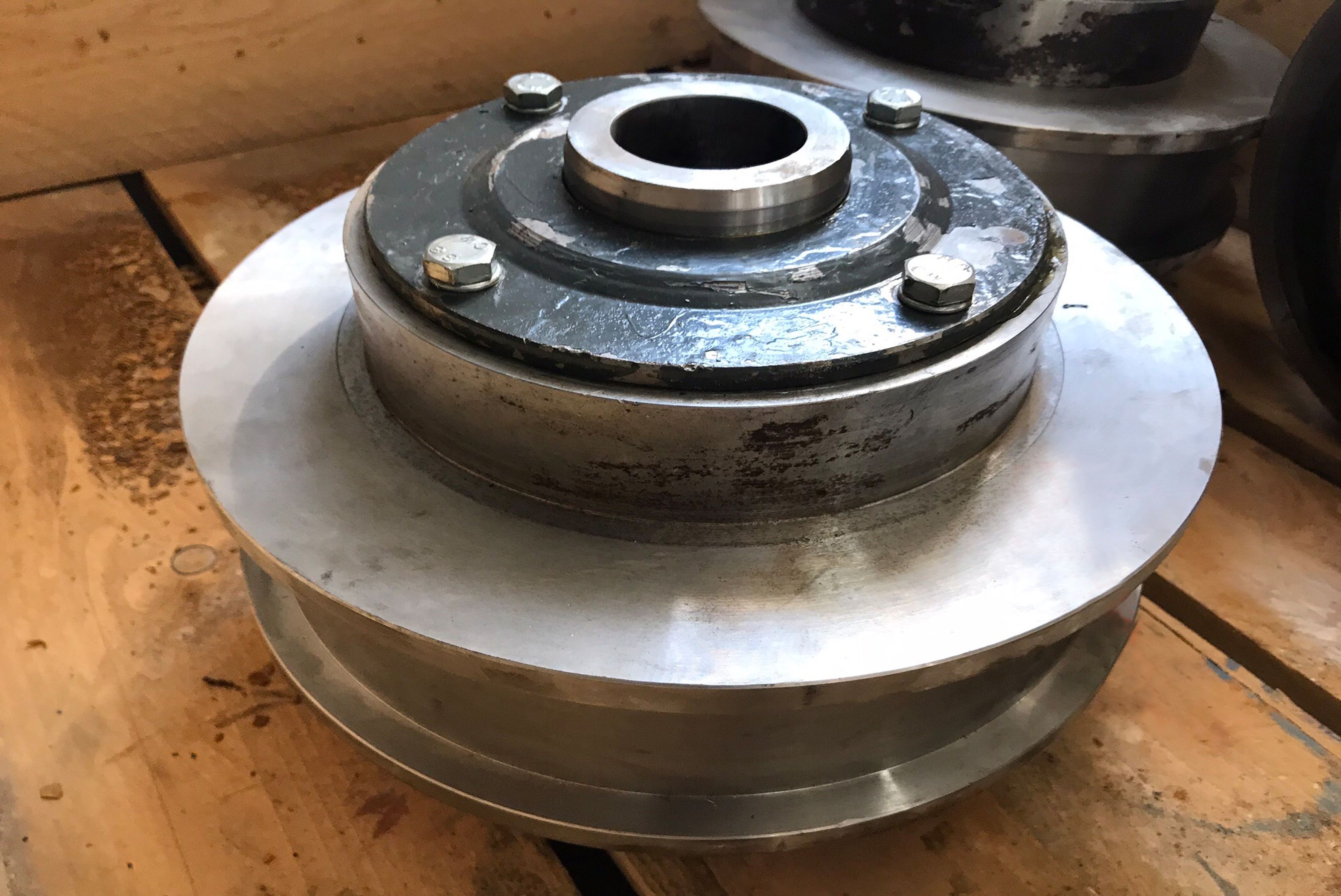 Un componente meccanico industriale in metallo lavorato, posizionato su un pallet di legno, caratterizzato da un asse centrale fissato da quattro viti di montaggio sulla parte superiore.