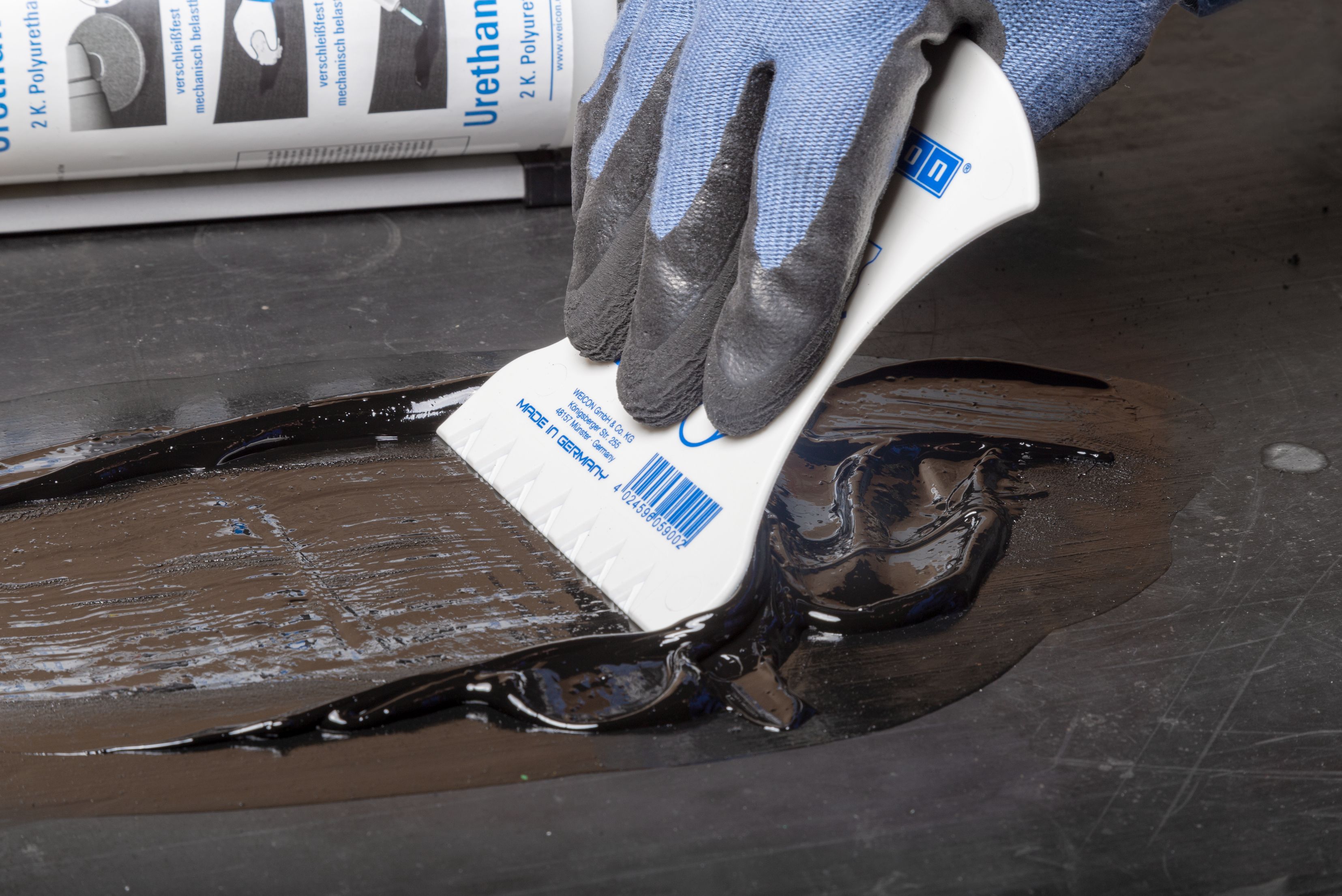 Una persona indossa guanti protettivi e utilizza una spatola di plastica bianca per stendere e livellare una resina di riparazione nera su una superficie danneggiata.