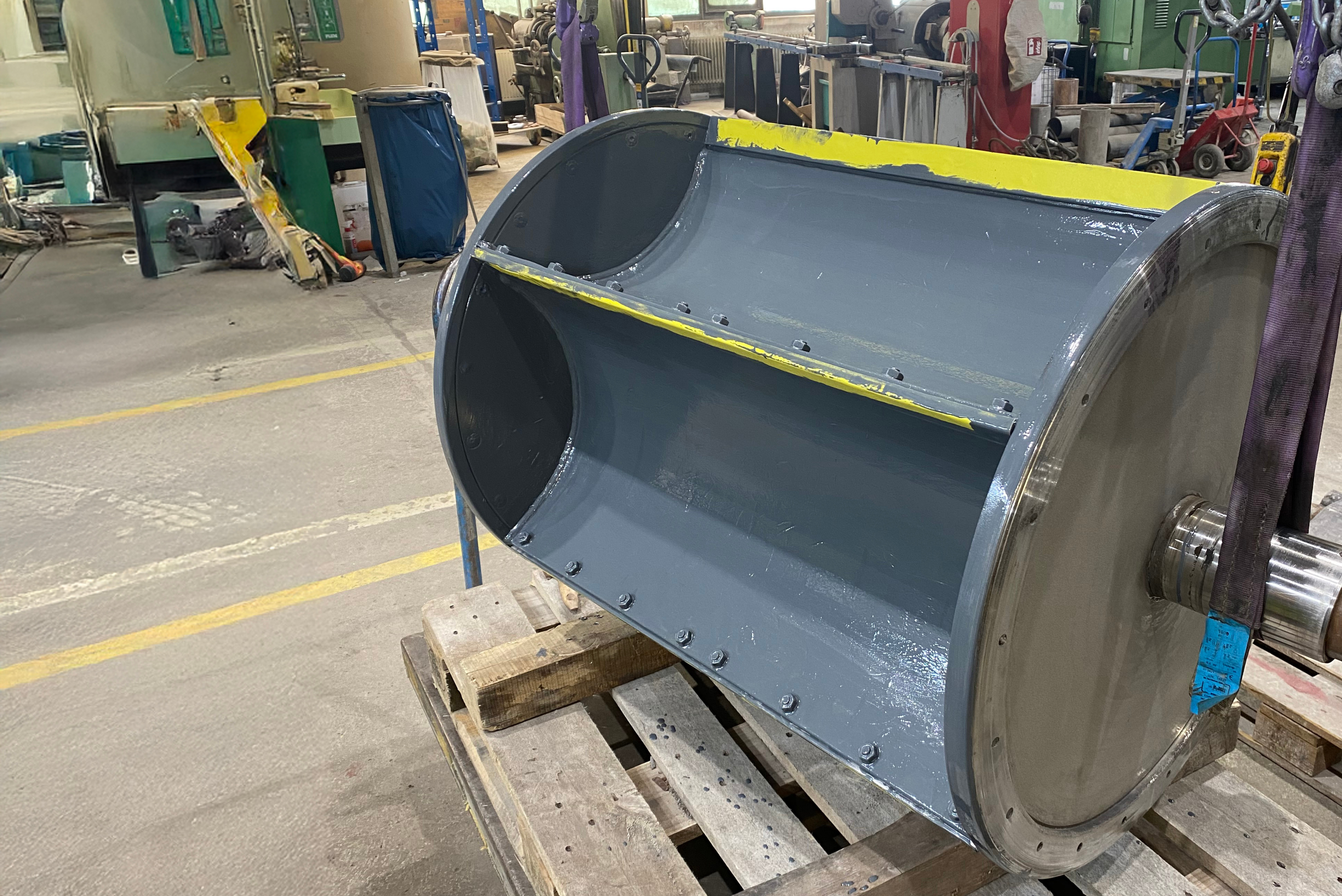 Un grande rullo o tamburo industriale rivestito con una vernice protettiva grigia, posizionato su pallet di legno all'interno di un'officina.