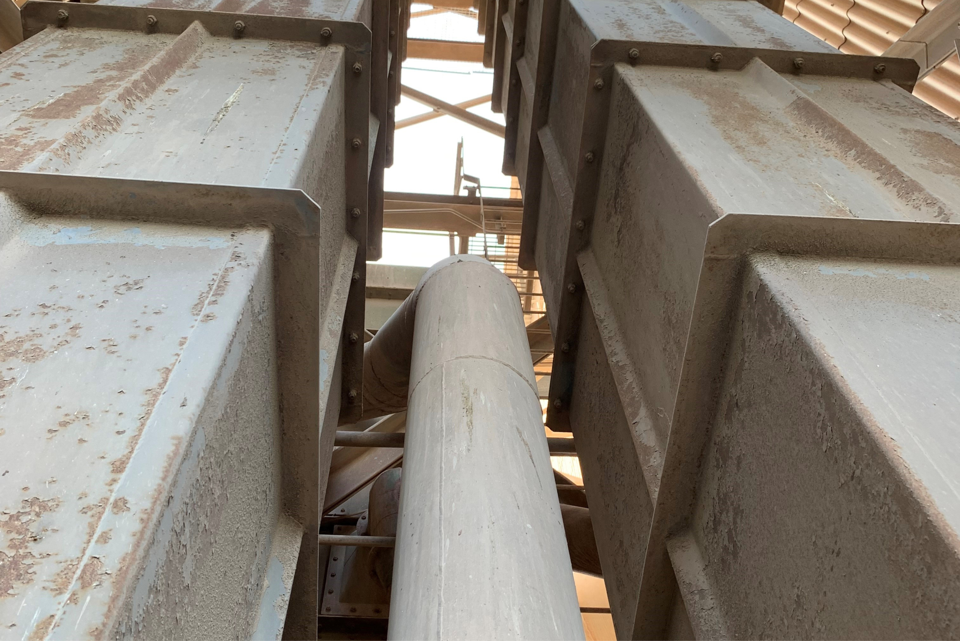 Una vista dal basso mostra due grandi condotti industriali verticali a sezione rettangolare, caratterizzati da superfici metalliche usurate e bordi flangiati bullonati, con un tubo cilindrico centrale che sale verticalmente tra di essi.