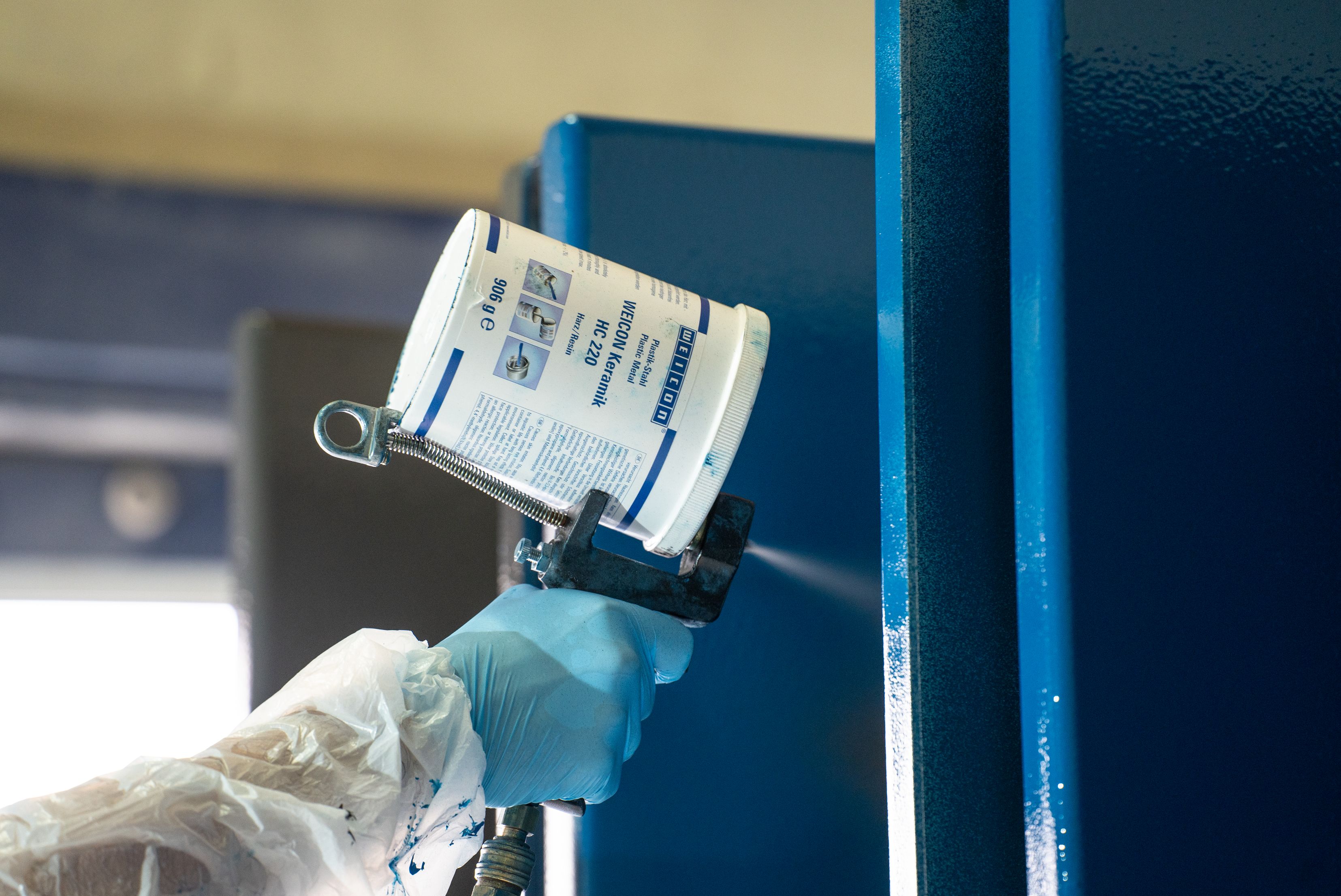 Un tecnico indossa guanti protettivi blu e abbigliamento protettivo mentre utilizza una pistola a spruzzo con un contenitore di rivestimento WEICON per applicare una vernice blu su una superficie metallica industriale.