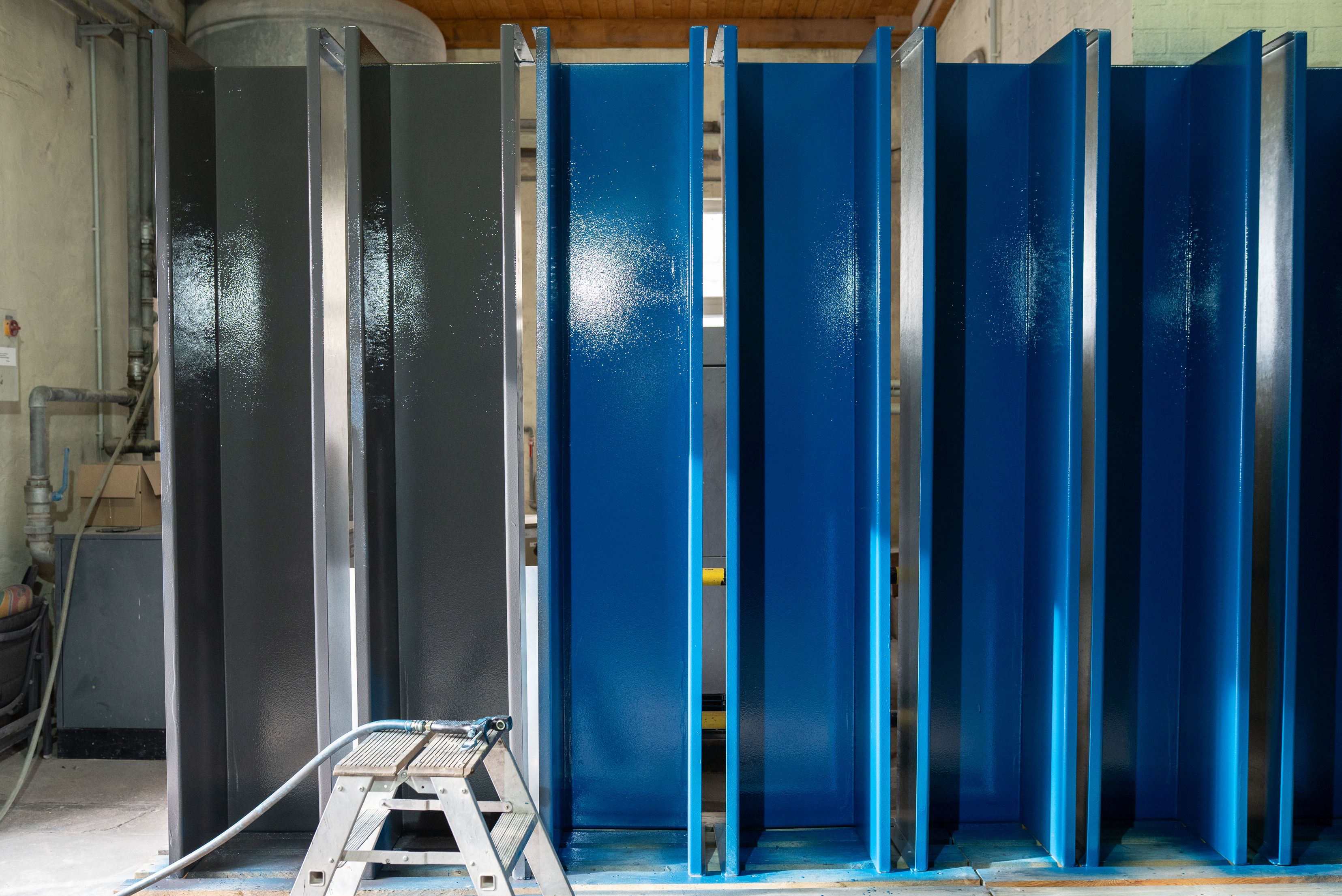 Una serie di grandi componenti metallici industriali verticali allineati in un'officina, con sezioni trattate in colore grigio a sinistra e una finitura protettiva lucida di colore blu brillante sulla parte destra.