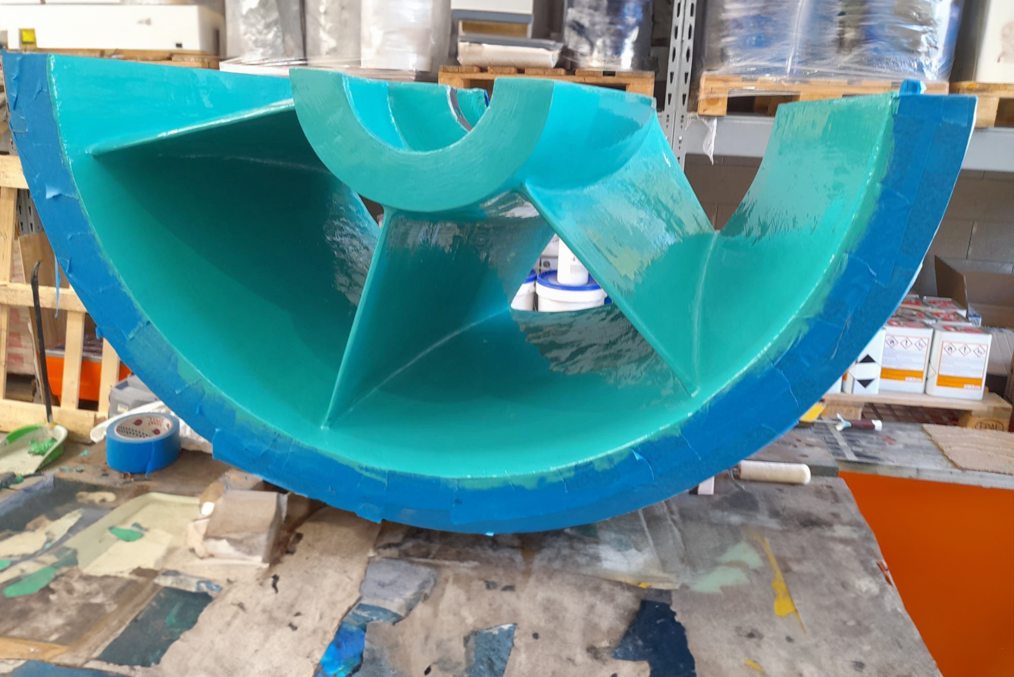 Una grande girante industriale semicircolare perfettamente rivestita con una protezione superficiale brillante di colore blu e verde, posizionata su un banco da lavoro all'interno di un'officina.