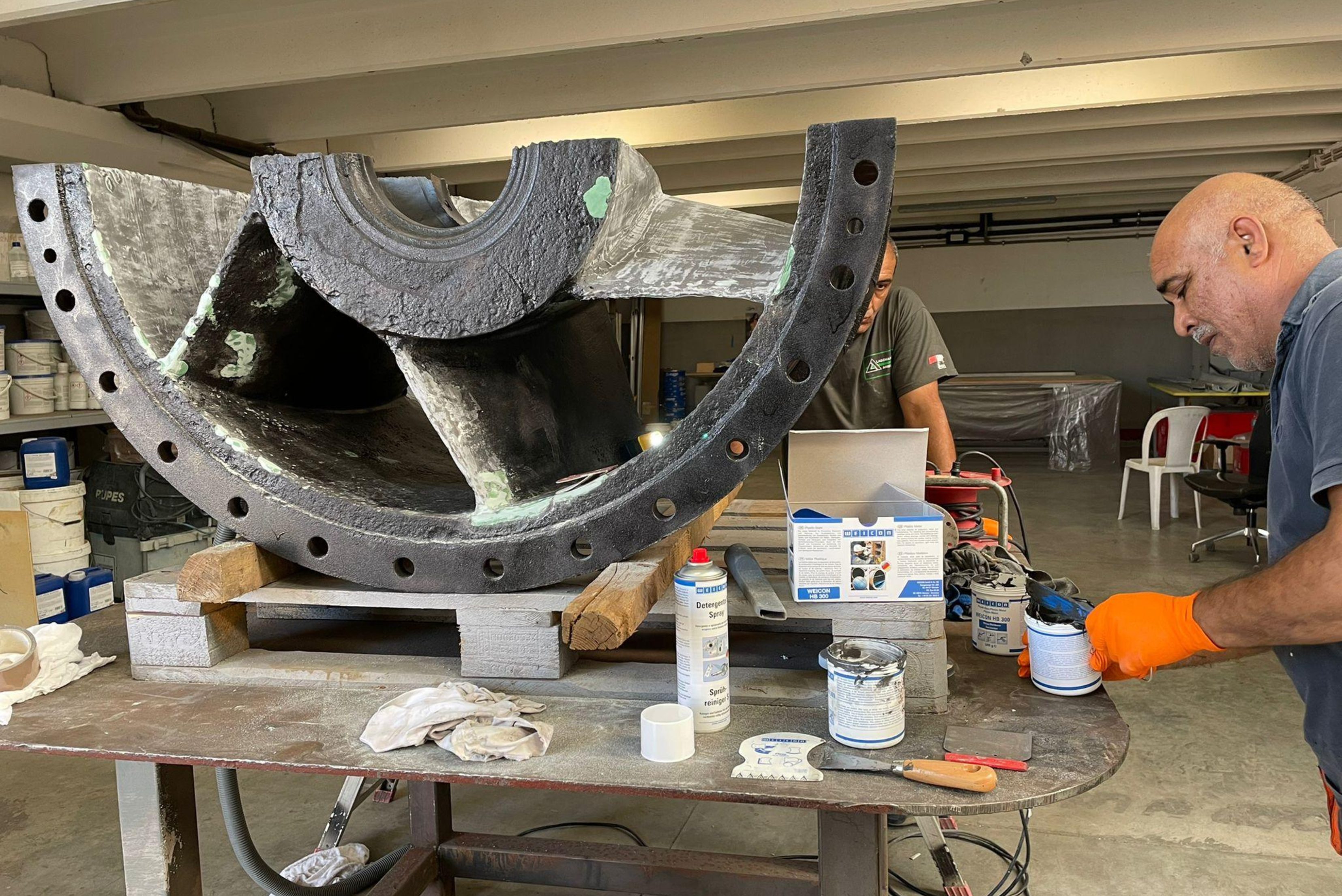 Un tecnico con guanti arancioni prepara materiali di rivestimento protettivo WEICON accanto a una grande girante industriale semicircolare posizionata su un banco da lavoro in officina.