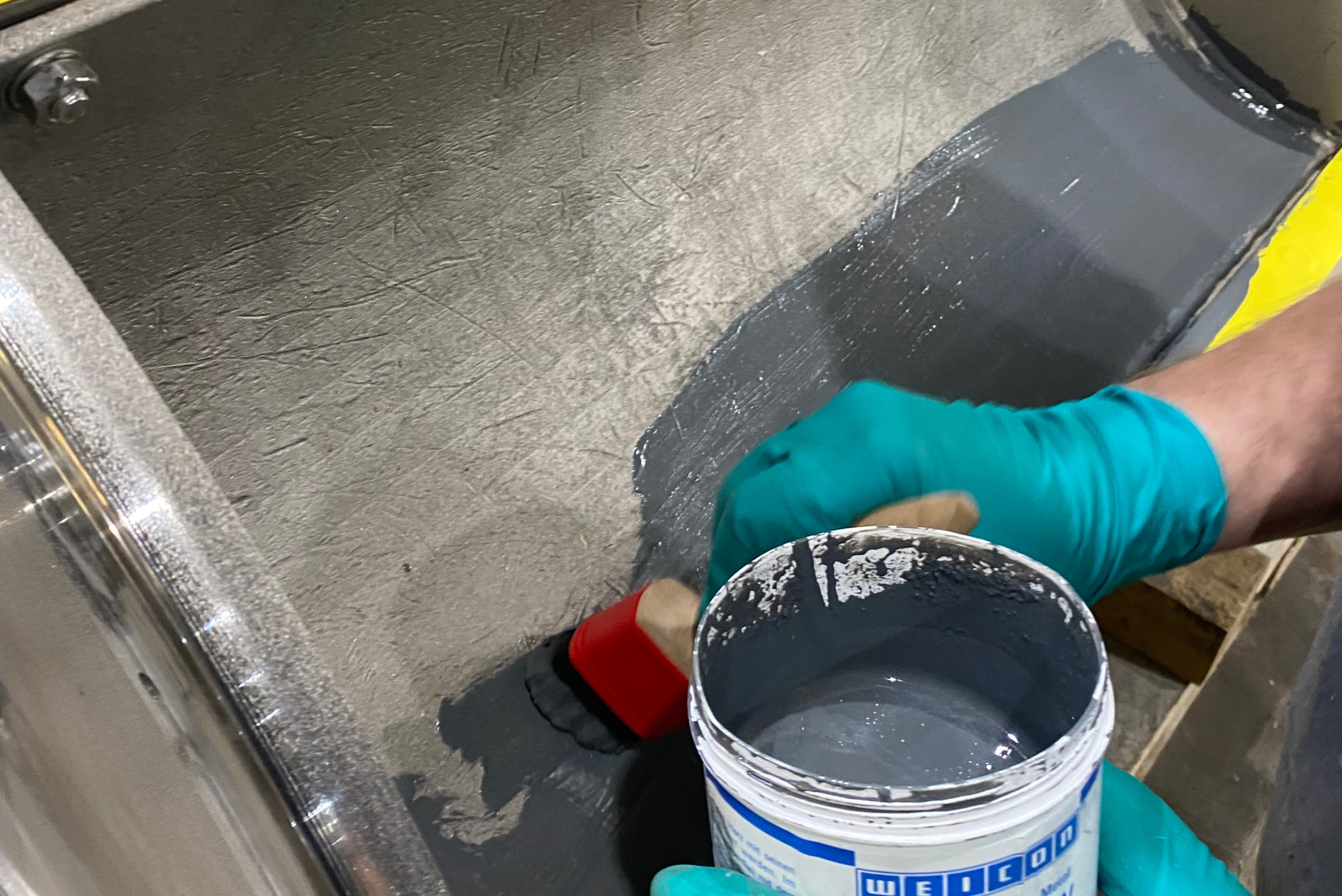 Una persona con guanti di protezione verdi applica un rivestimento protettivo grigio con un pennello sulla superficie interna di un componente industriale, tenendo in mano un barattolo del marchio WEICON.