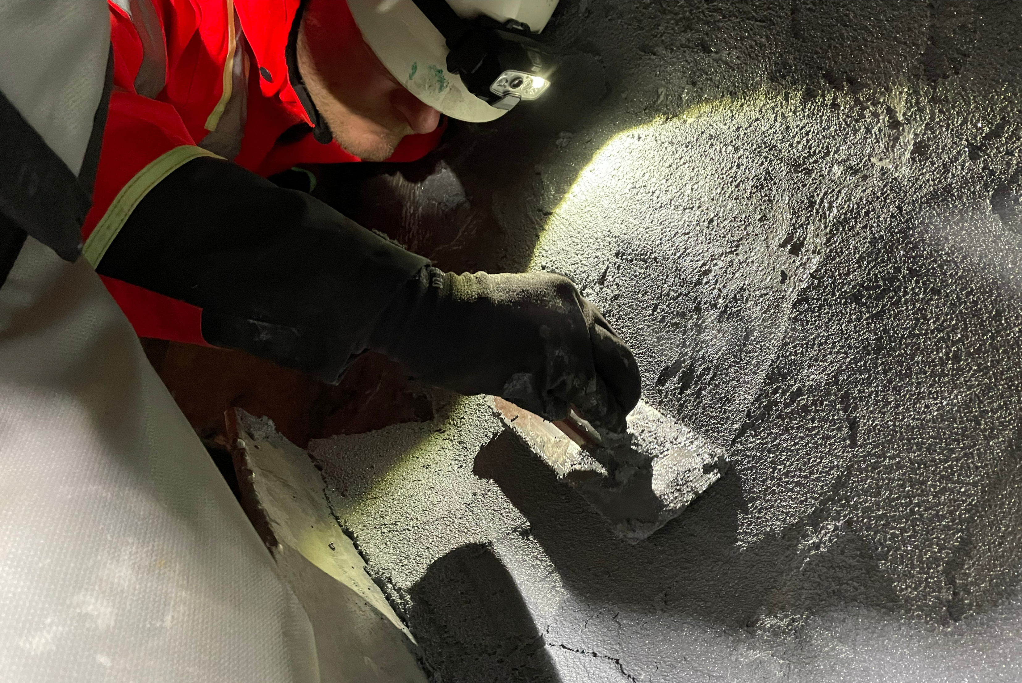 Un tecnico in abbigliamento protettivo con elmetto e lampada frontale applica malta di riparazione con una cazzuola su una superficie metallica irregolare e riparata.