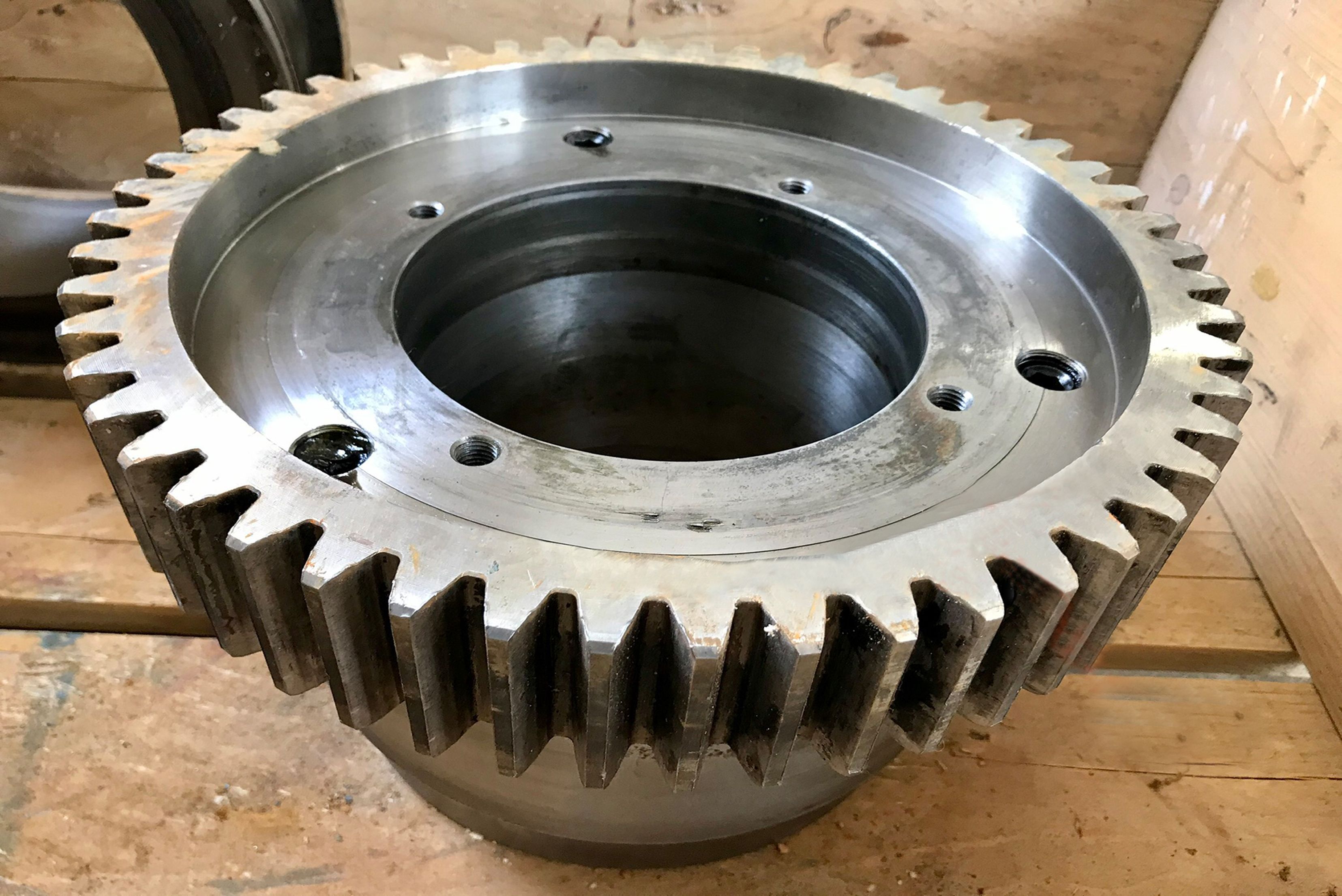 Un grande ingranaggio cilindrico industriale in metallo con denti esterni lavorati con precisione e fori di montaggio filettati sulla superficie superiore, posizionato su una superficie di legno.