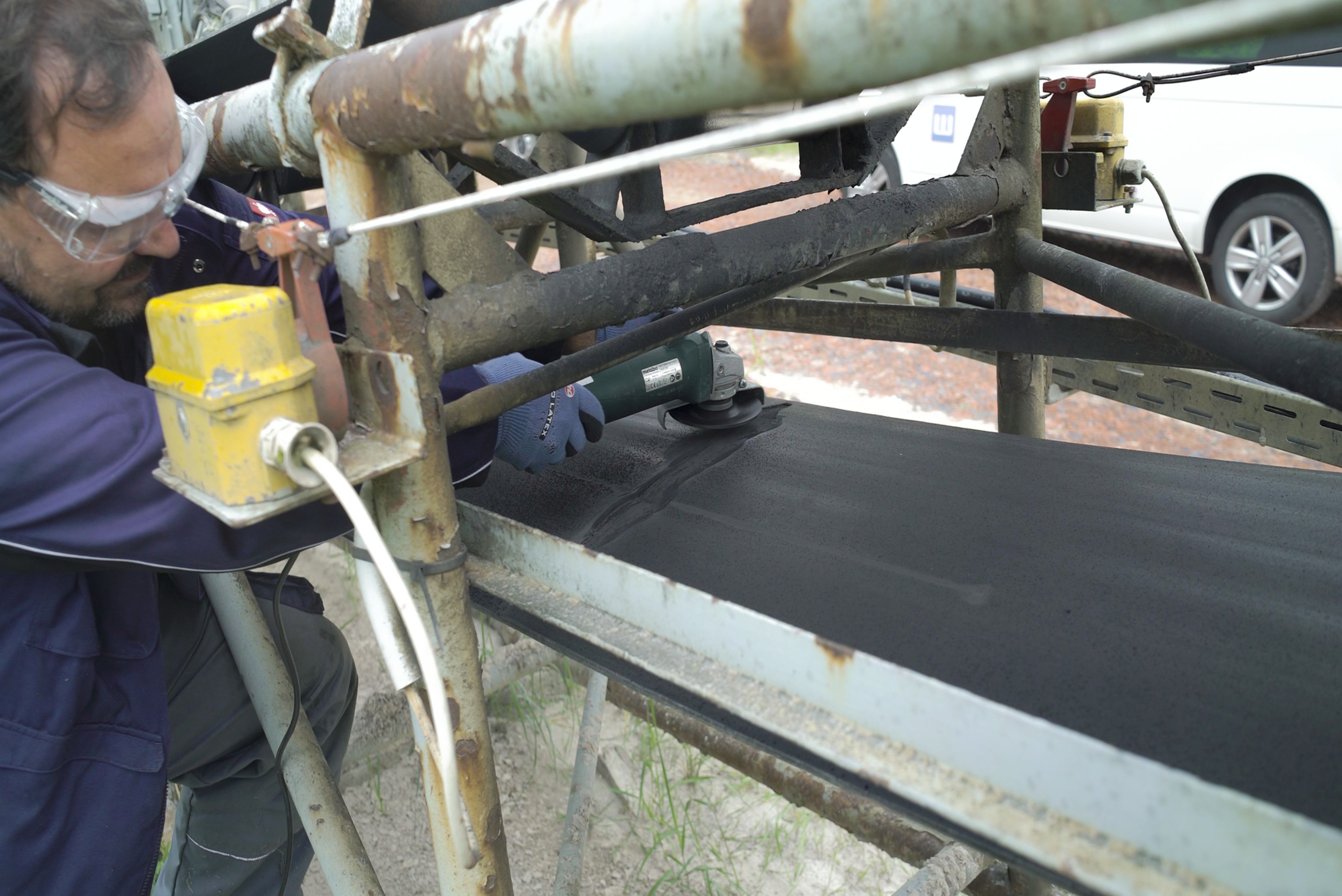 Un operaio con occhiali di protezione e guanti lavora su un nastro trasportatore industriale utilizzando una smerigliatrice angolare.