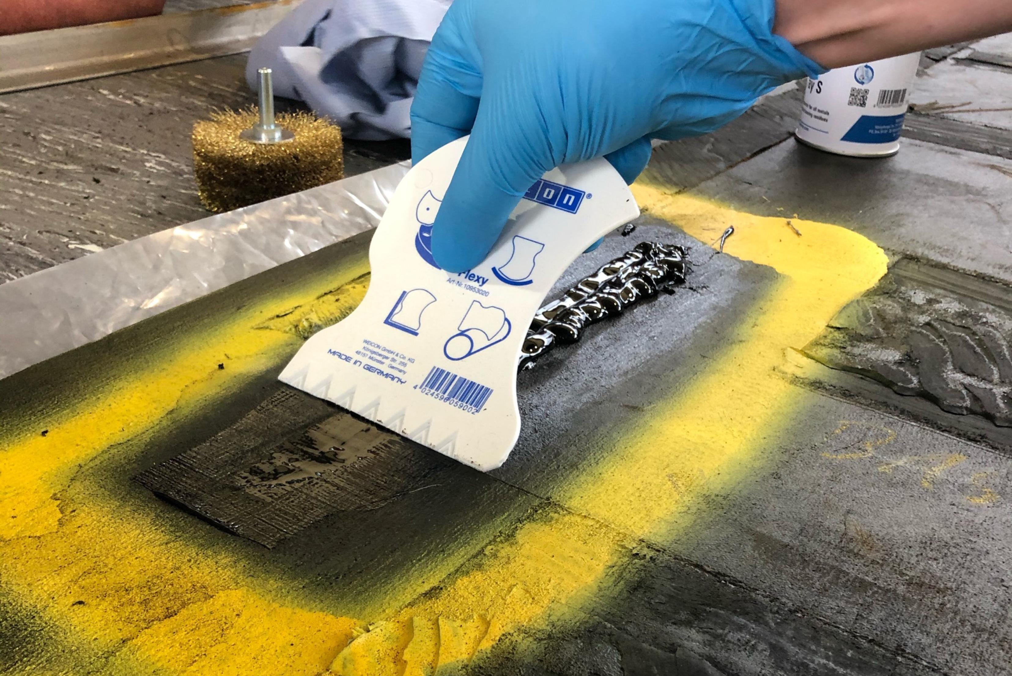 Una persona con guanti azzurri usa una spatola di plastica bianca per livellare la resina di riparazione nera su una superficie riparata.
