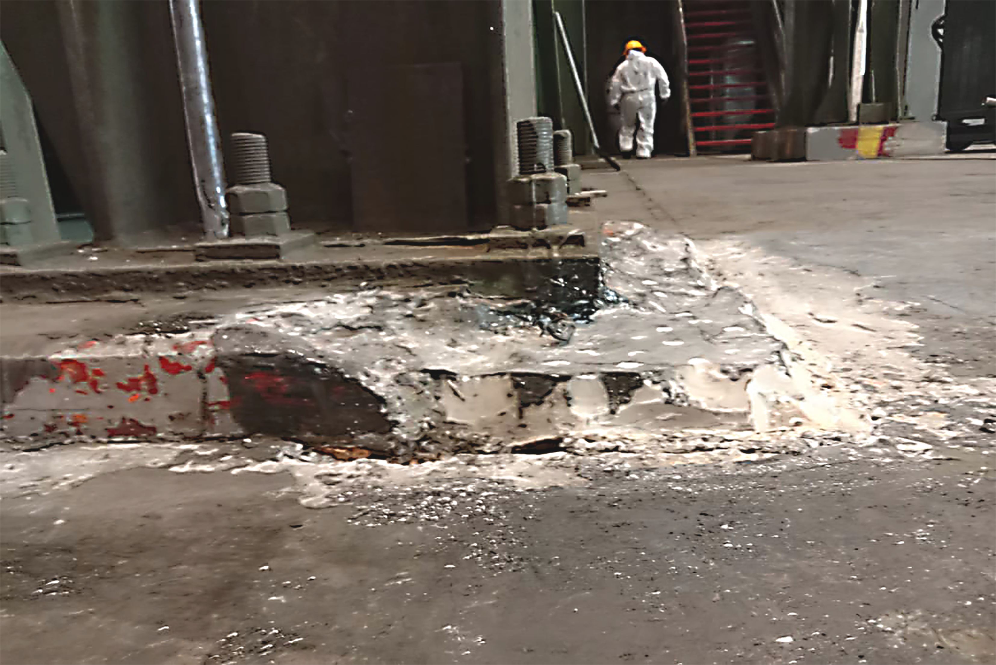 Base di macchinario industriale in calcestruzzo danneggiata con bulloni d'ancoraggio a vista e un operaio in tuta protettiva sullo sfondo.