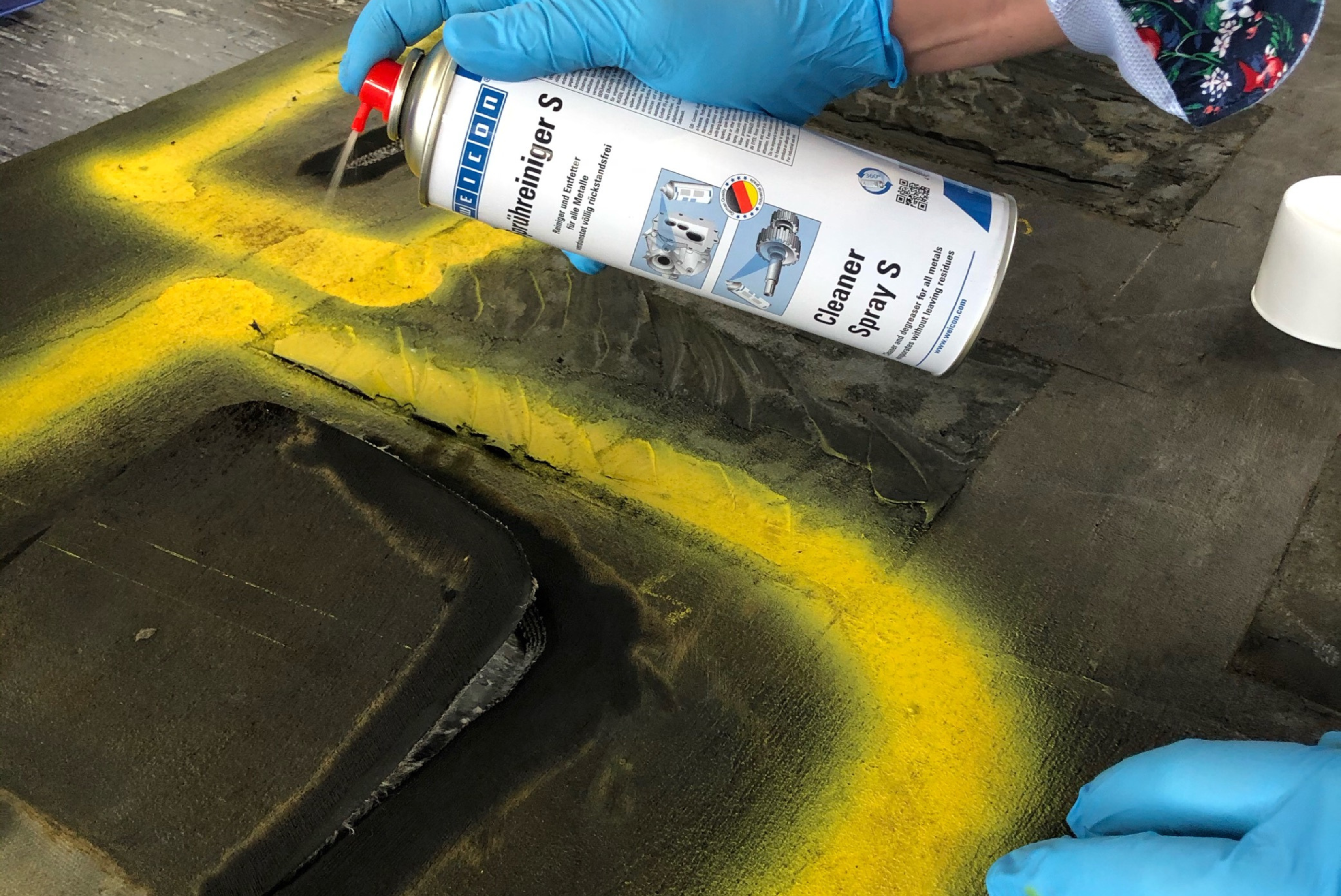 Una persona con guanti azzurri applica uno spray detergente industriale su una superficie metallica contrassegnata da linee gialle.