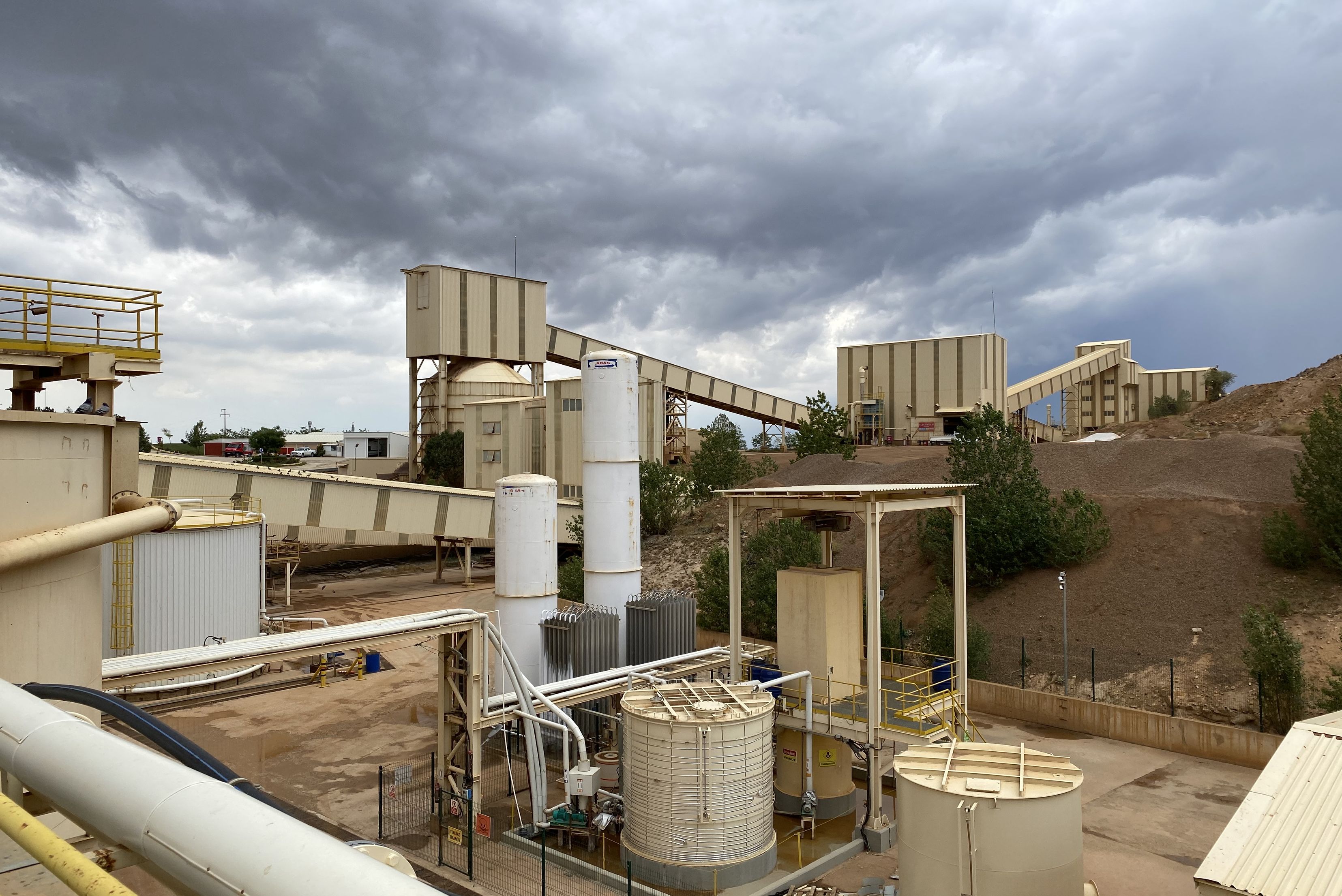 Panoramica esterna di un grande impianto industriale con silos, serbatoi di stoccaggio e nastri trasportatori coperti sotto un cielo nuvoloso.